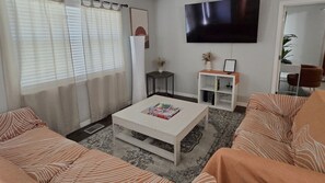 A living room with a white coffee table, two sofas with striped cushions, a flat-screen TV on the wall, and a small side table with a vase and picture frame. - Comfort Cove, 3BR, 2BA, Fully Fenced, Spring Hill, FL (Spring Hill)