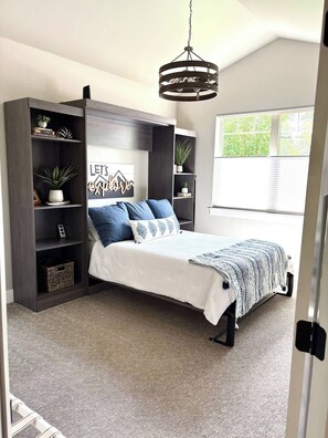 A bedroom with a bed, dark wooden shelving, a window, and a hanging light fixture.