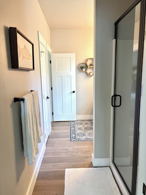 Un pasillo con piso de madera, una puerta blanca y una puerta de cristal que da a un baño.