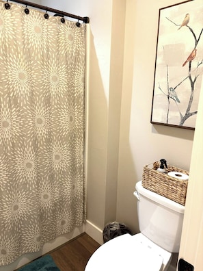 Une salle de bain avec un rideau de douche, des toilettes, une poubelle et un tableau encadré au mur.