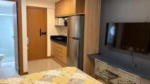 A compact kitchen with wooden cabinets, a refrigerator, a microwave, and a flat-screen TV mounted on the wall.