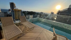 A rooftop pool with lounge chairs and a glass balustrade overlooking a cityscape.