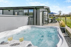 Un bain à remous extérieur moderne, avec deux verres de vin blanc, un espace patio aménagé avec du mobilier d’extérieur et une vue sur un paysage verdoyant.
