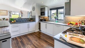 A modern kitchen with white cabinets, a wooden floor, and a large window providing natural light. - 3 Bed in Broadwoodwidger (oc-h31771) (Broadwoodwidger)