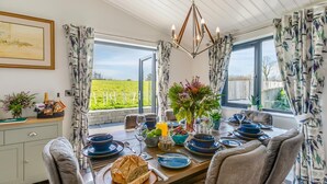 A dining area with a table set for four, featuring a chandelier, floral arrangements, and a view of the outdoors. - 3 Bed in Broadwoodwidger (oc-h31771) (Broadwoodwidger)