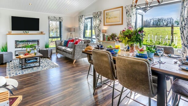 A spacious living room with a fireplace, a dining area with a table set for two, and a large window offering a view of the outdoors. - 3 Bed in Broadwoodwidger (oc-h31771) (Broadwoodwidger)