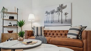 Un salón con un sofá de cuero marrón, dos mesas de centro de mármol blanco, una lámpara de pie, una estantería con objetos decorativos y un cuadro con motivos playeros.