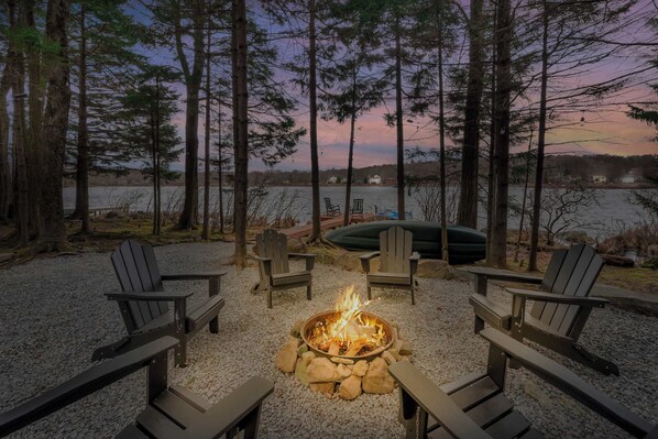 A fire pit surrounded by Adirondack chairs, a lake view, and a dock with a boat.