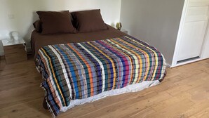 A bedroom with a wooden floor, a bed with a colorful striped blanket, two brown pillows, a white nightstand with a lamp, and a white door. - Room at the foot of the Rhune (Sare)