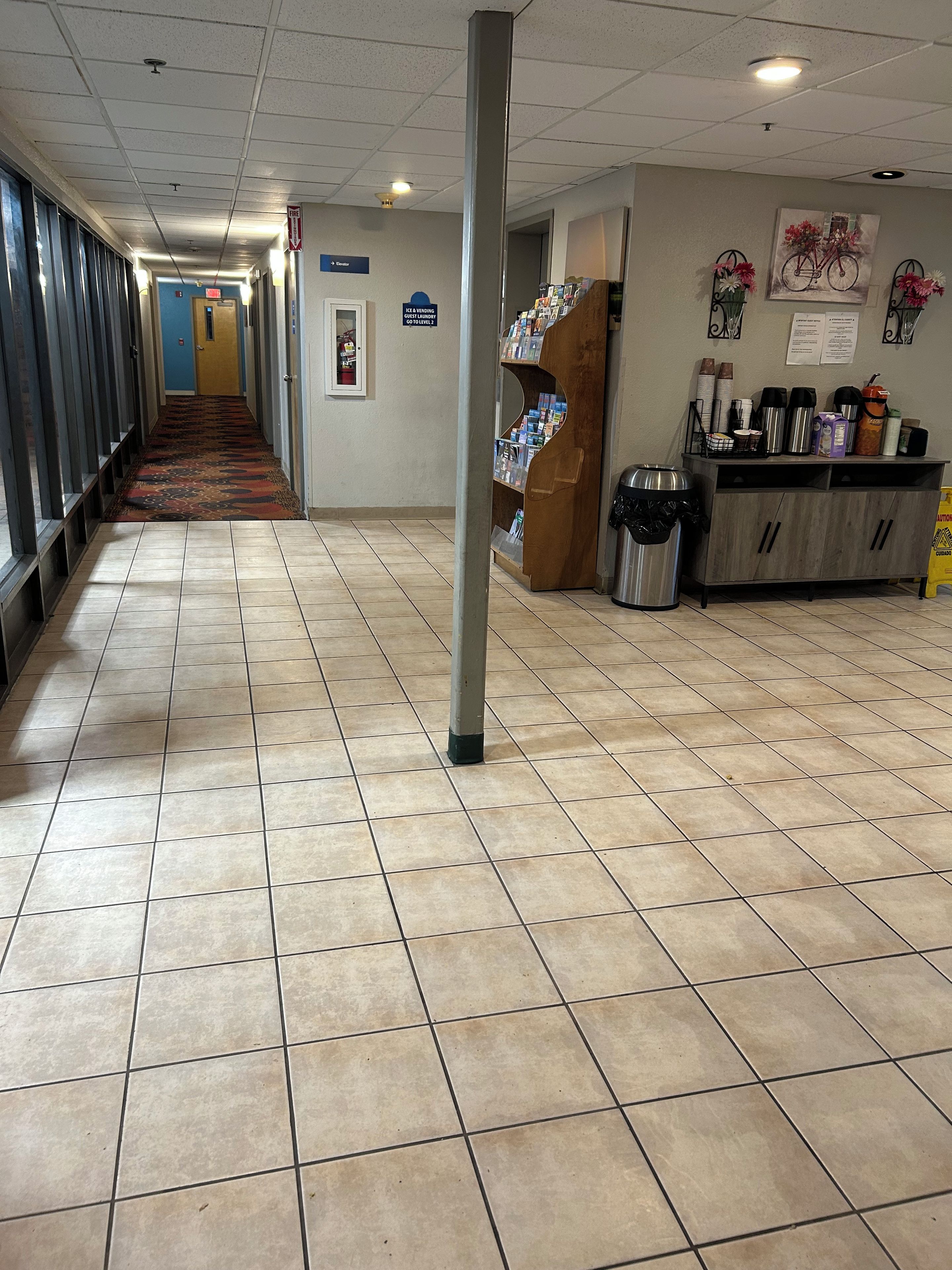 A hotel corridor with a tiled floor, a vending machine, a bulletin board, and a coffee station.