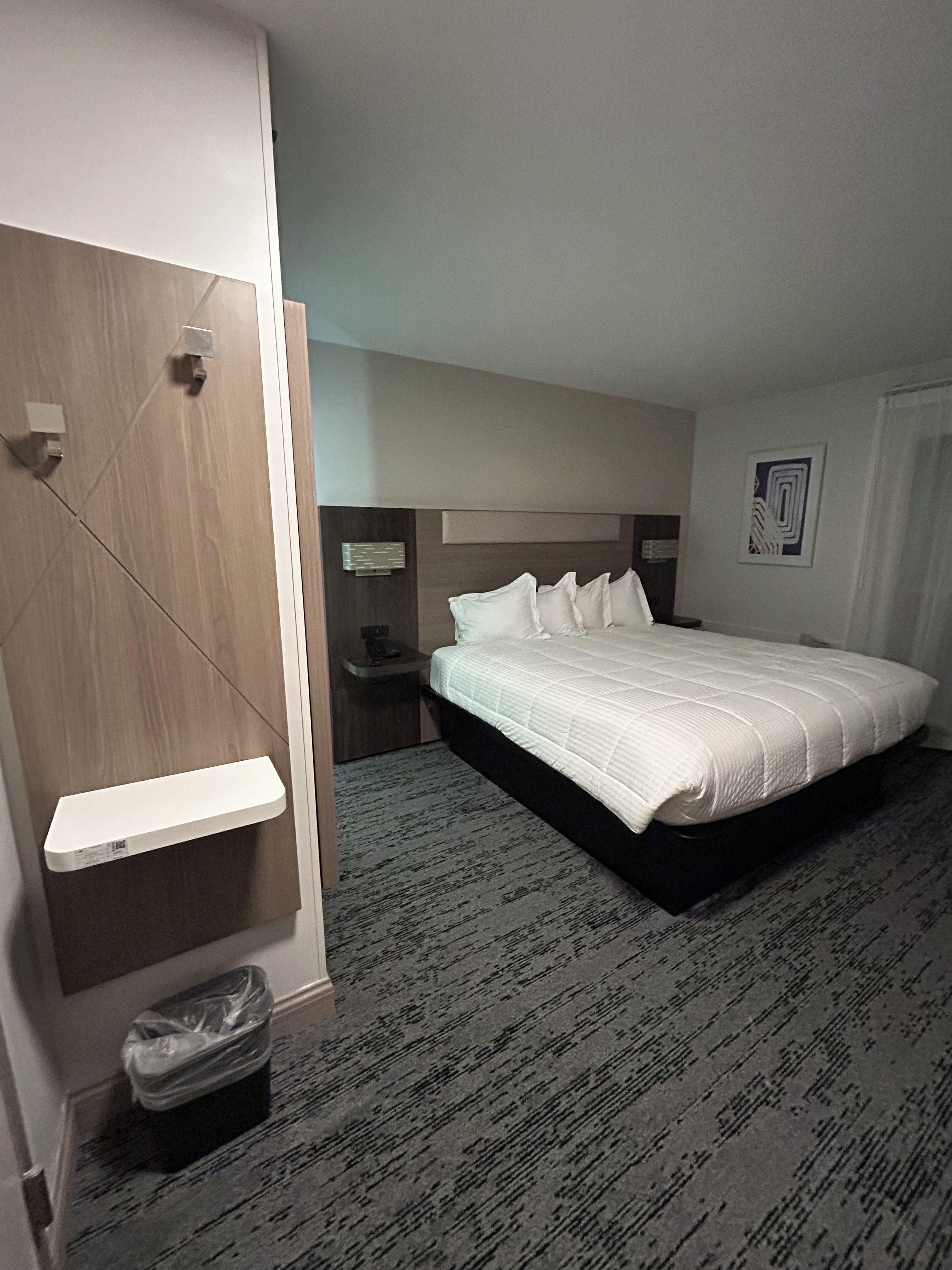 Een hotelkamer met een bed, nachtkastje, prullenbak en een wandplank.