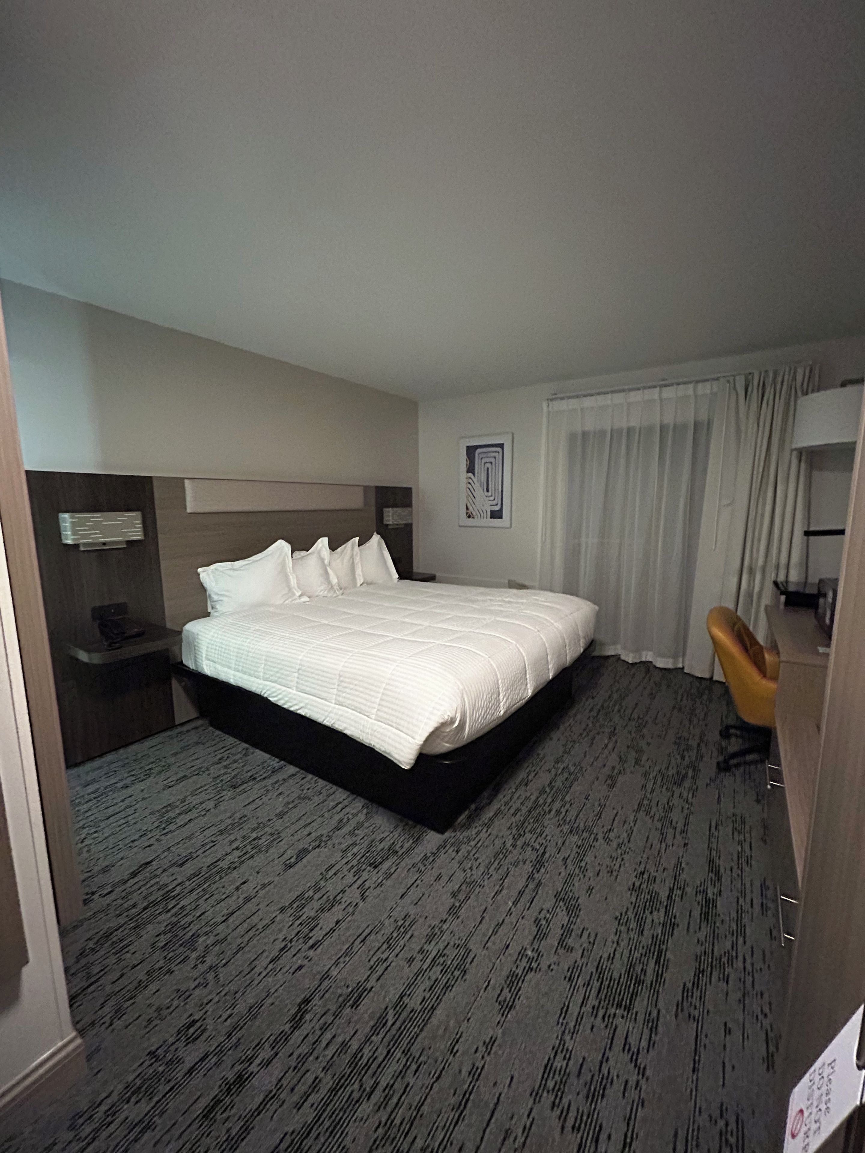 Una habitación de hotel con una cama grande, un escritorio, una silla y un ventanal con cortinas.