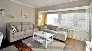 A living room with a sofa, a coffee table, and a window with a view of buildings. - Apartment in Knokke Near Albertstrand Beach (Knokke-Heist)