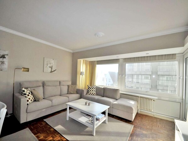 A living room with a sofa, a coffee table, and a window with a view of buildings. - Apartment in Knokke Near Albertstrand Beach (Knokke-Heist)