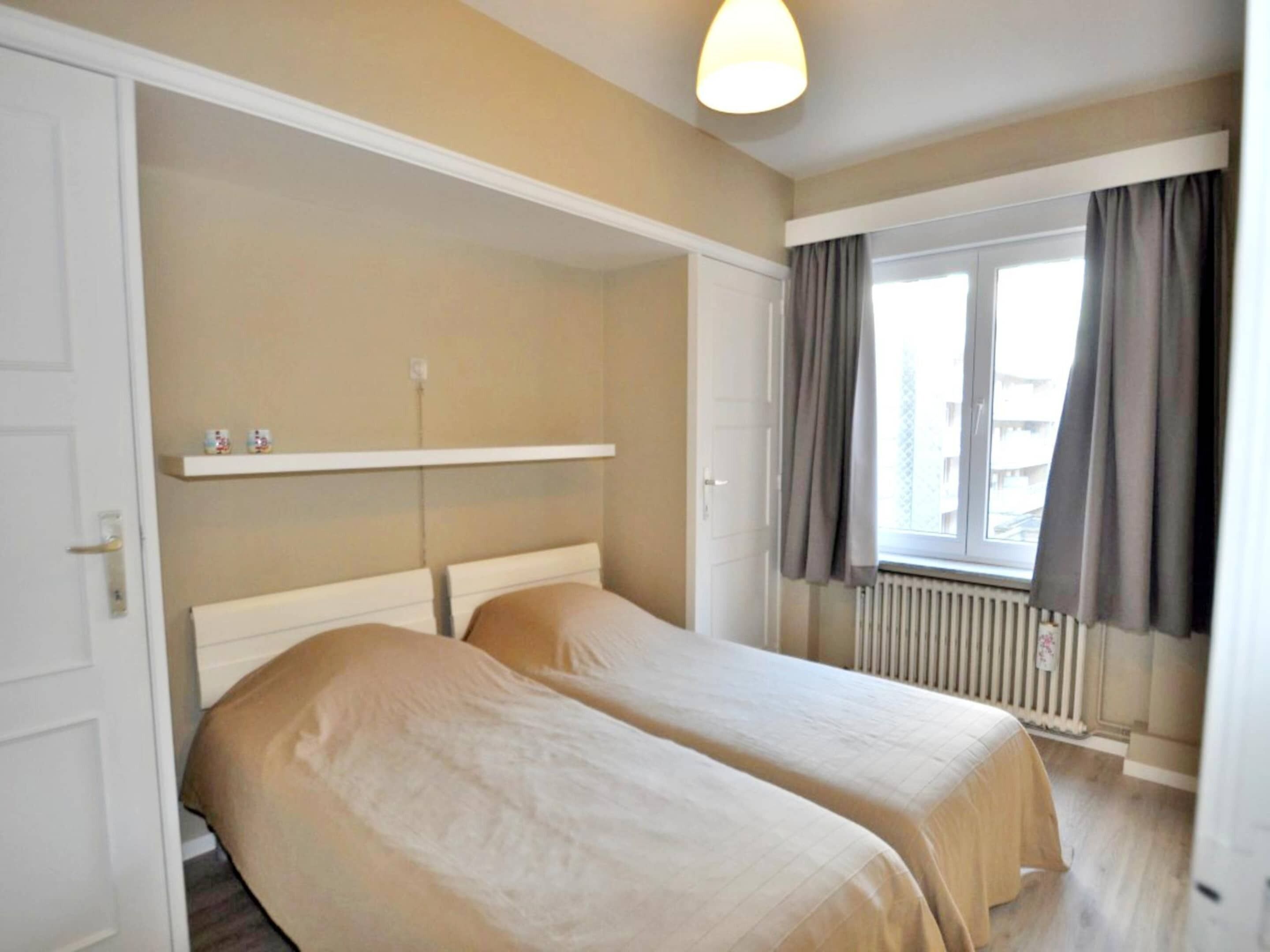 A room with two beds, a window with curtains, a radiator, and a shelf with items.