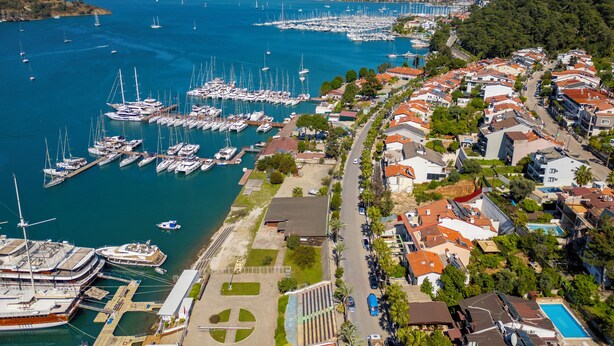An aerial view of a marina with numerous boats docked, surrounded by residential buildings with red-tiled roofs and a mountainous backdrop. - Cennet Life Boutique Hotel (Fethiye)