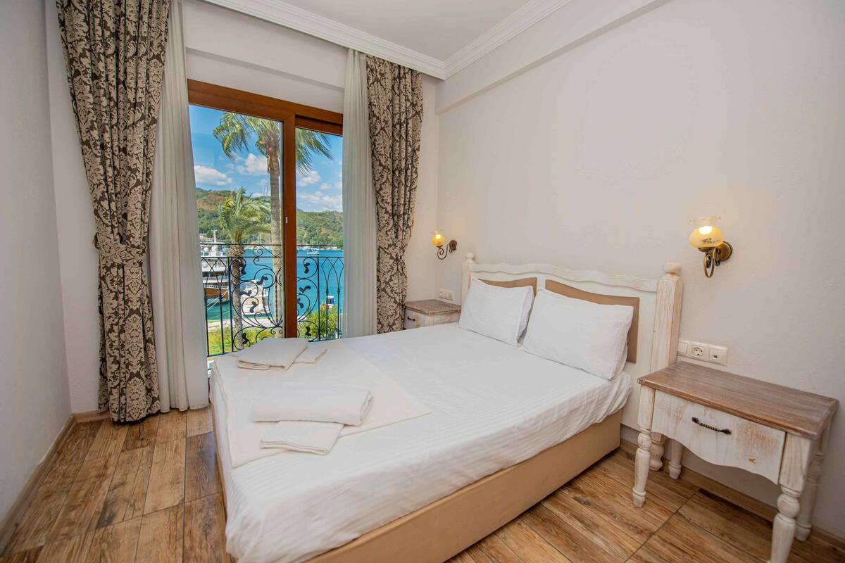 A bedroom with a large bed, a wooden nightstand, and a view of a pool and palm trees through a window.