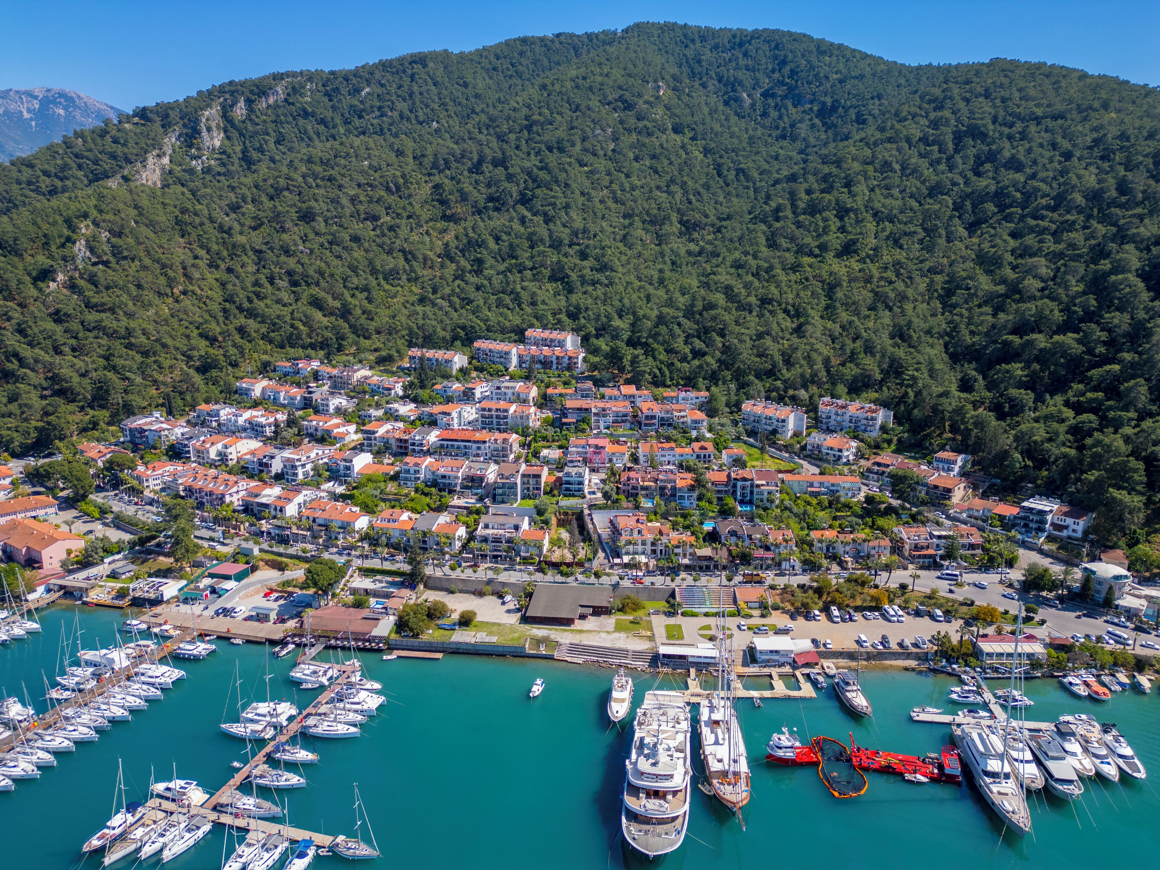 Uma vila costeira com uma marina repleta de iates e barcos, cercada por colinas arborizadas.