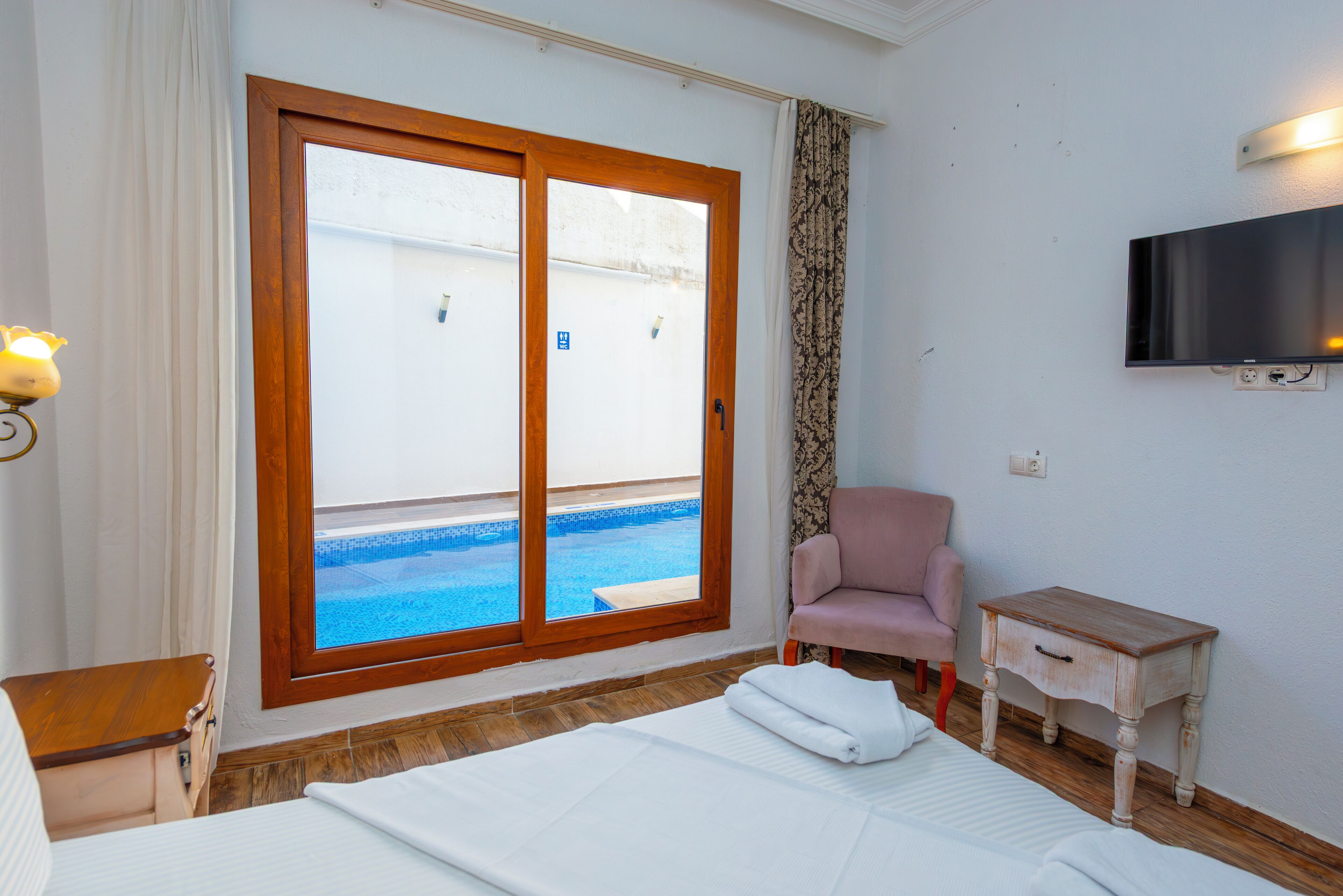 A hotel room with a bed, a chair, a small table, a TV, and a sliding glass door leading to a pool.