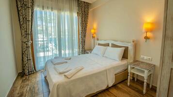 Une chambre d’hôtel avec un grand lit, deux lampes de chevet, une petite table de nuit en bois et une vue sur l’extérieur à travers des rideaux légers.