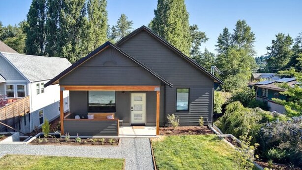 Een modern huis met twee verdiepingen, een overdekte veranda, een verzorgd gazon en een bebost achtergrond.