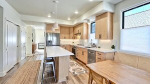 Een moderne keuken met een centraal eiland, houten kasten, roestvrijstalen apparatuur en een groot raam met jaloezieën.