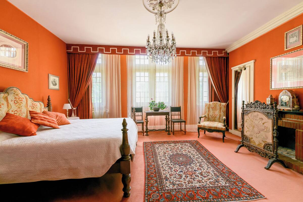 A room with a bed, a table with chairs, a chandelier, and a fireplace.