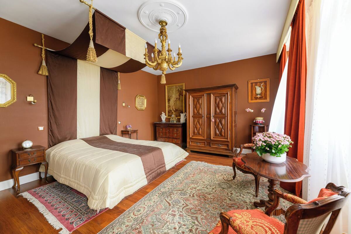 A bedroom with a large bed, a wooden wardrobe, a chandelier, and a table with a vase of flowers.