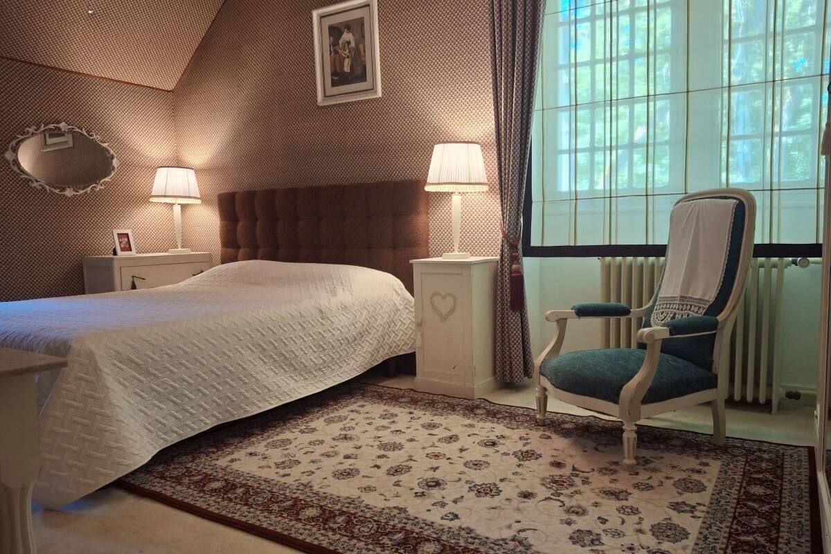 A bedroom with a bed, nightstands, a chair, a window with curtains, and a radiator.