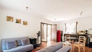 Una sala de estar con un sofá, un banco y un comedor con sillas y una mesa. Hay un ventilador, un calentador pequeño y un refrigerador en la zona de la cocina.