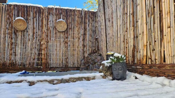 Una cerca de bambú con aberturas circulares, un muro de piedra y una estatua de Buda en un espacio exterior nevado.