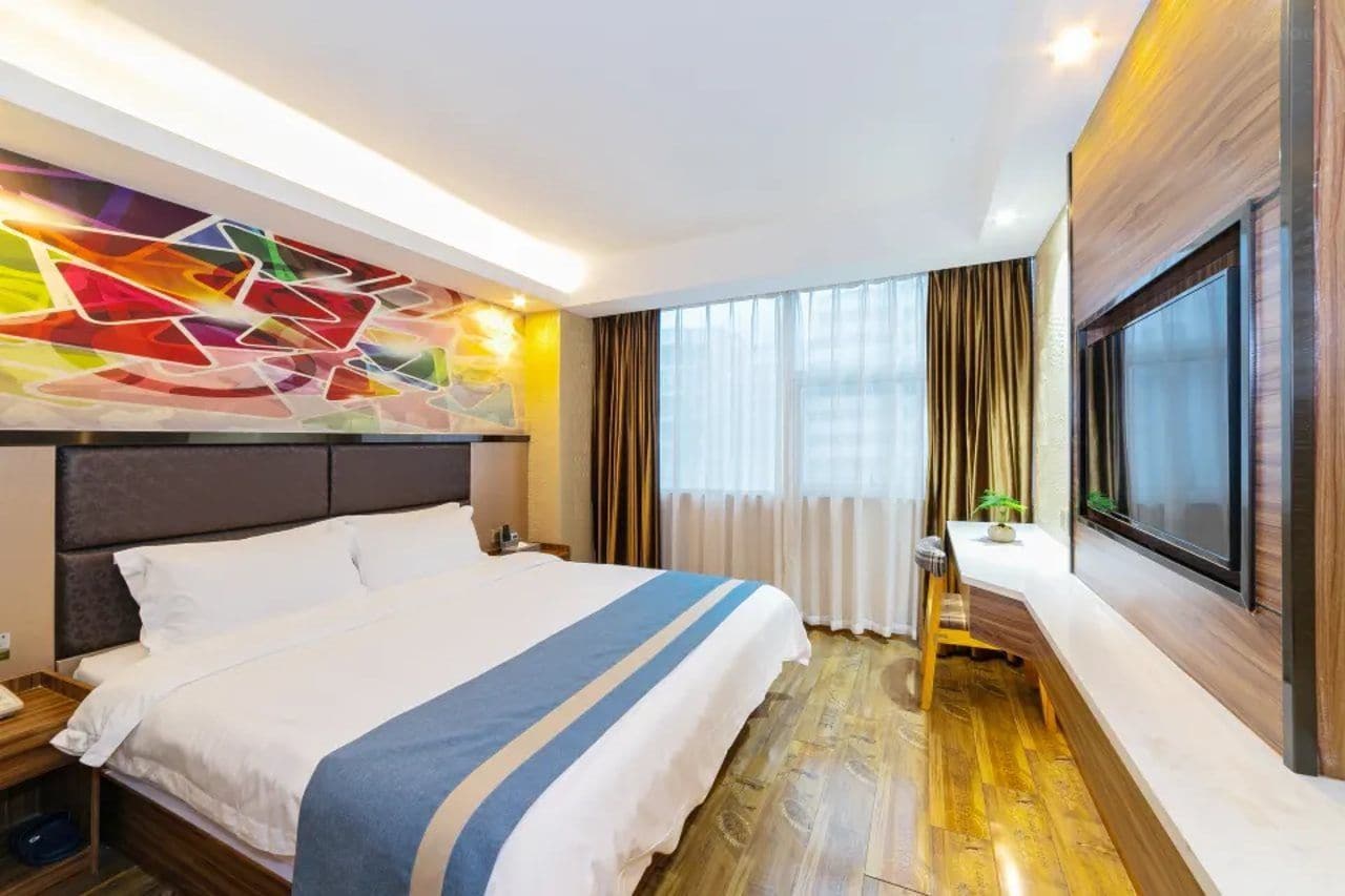 A modern hotel room with a large bed, a colorful abstract wall art, a large window with curtains, and a wooden floor.