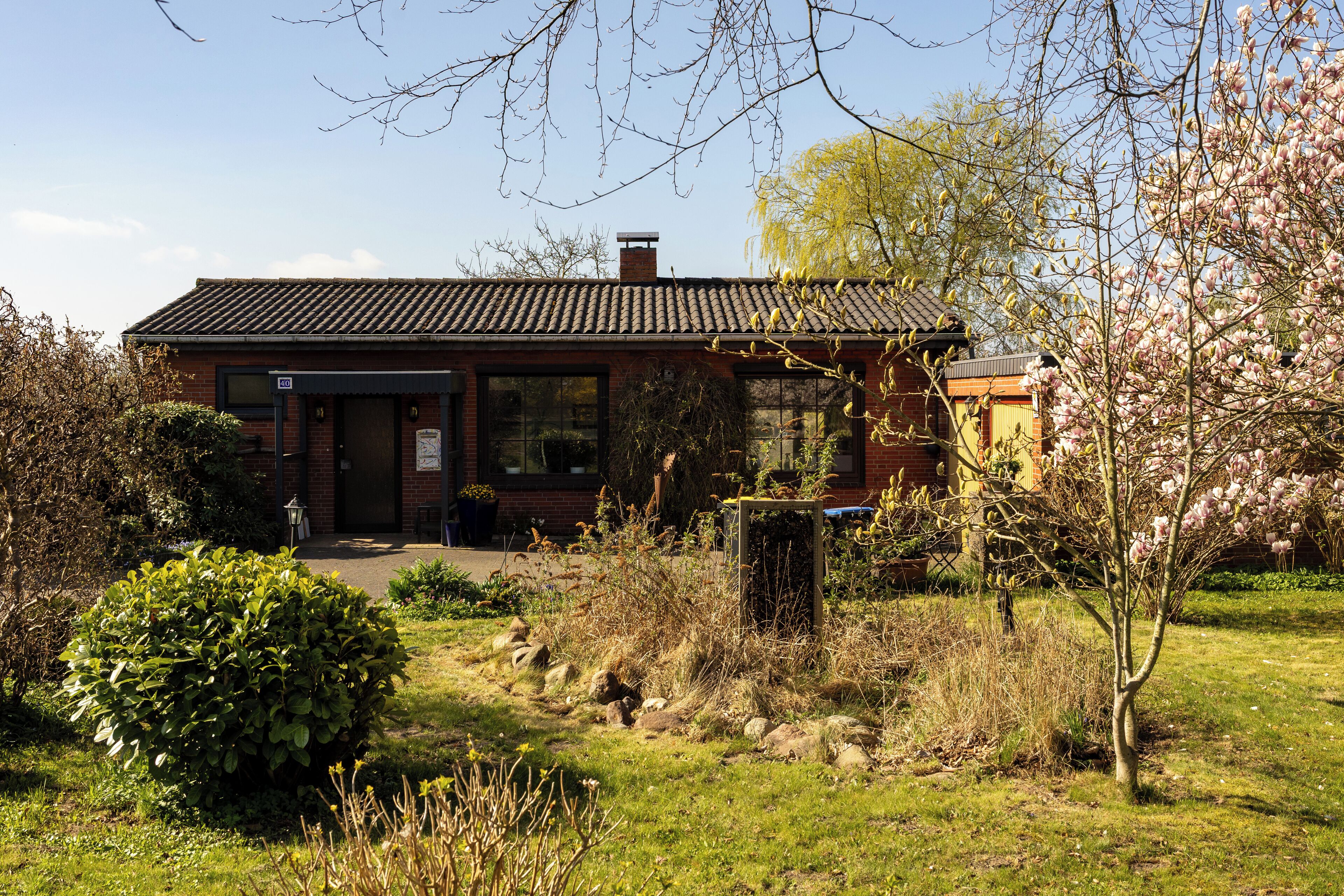 A brick building with a tiled roof, large windows, and a small garden area with bushes and a tree.