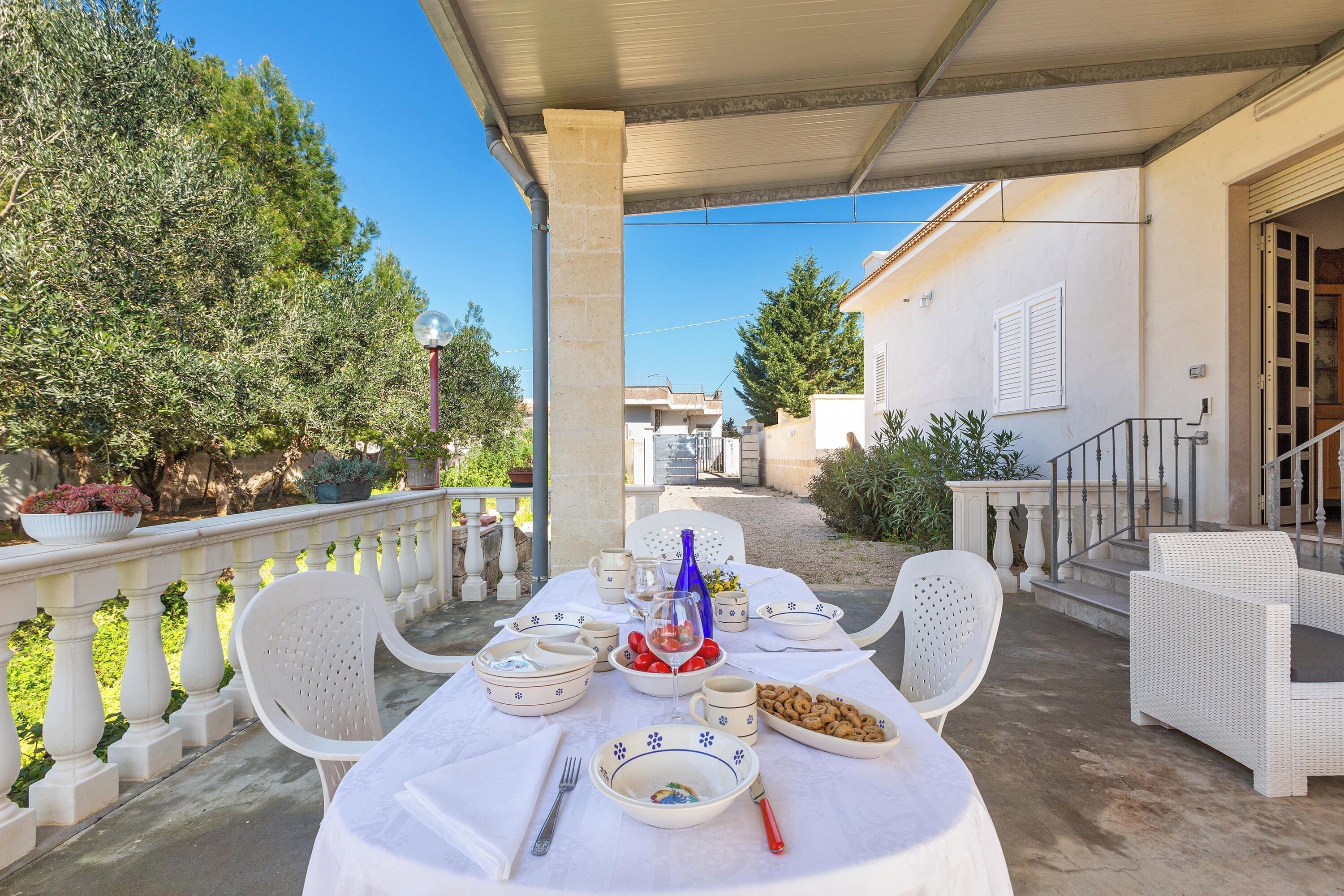 Un espace de restauration en plein air avec une table dressée pour quatre personnes, entourée de chaises en osier blanc, sous une terrasse couverte.