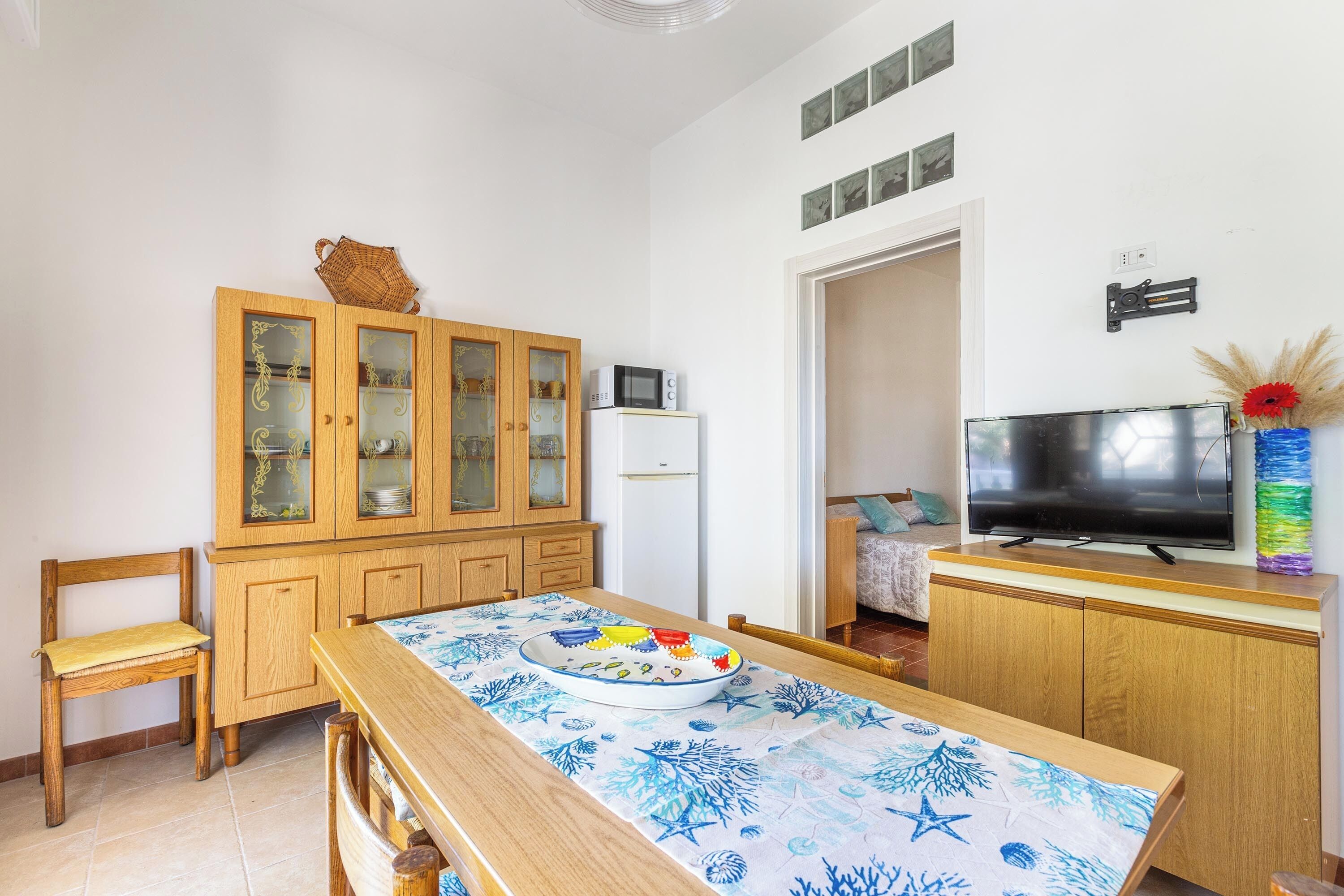 Ein kompakter Wohnraum mit Esstisch, Stühlen, einer Schrank mit Glastüren, einer Mikrowelle, einem Kühlschrank, einem Fernseher und einer Tür zu einem weiteren Raum.