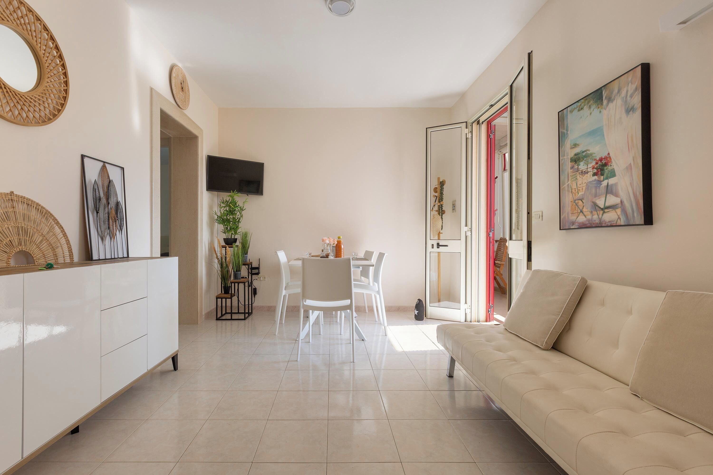 Una sala de estar moderna con un sofá, una mesa de comedor y sillas. Hay un televisor montado en la pared y un cuadro sobre el sofá.