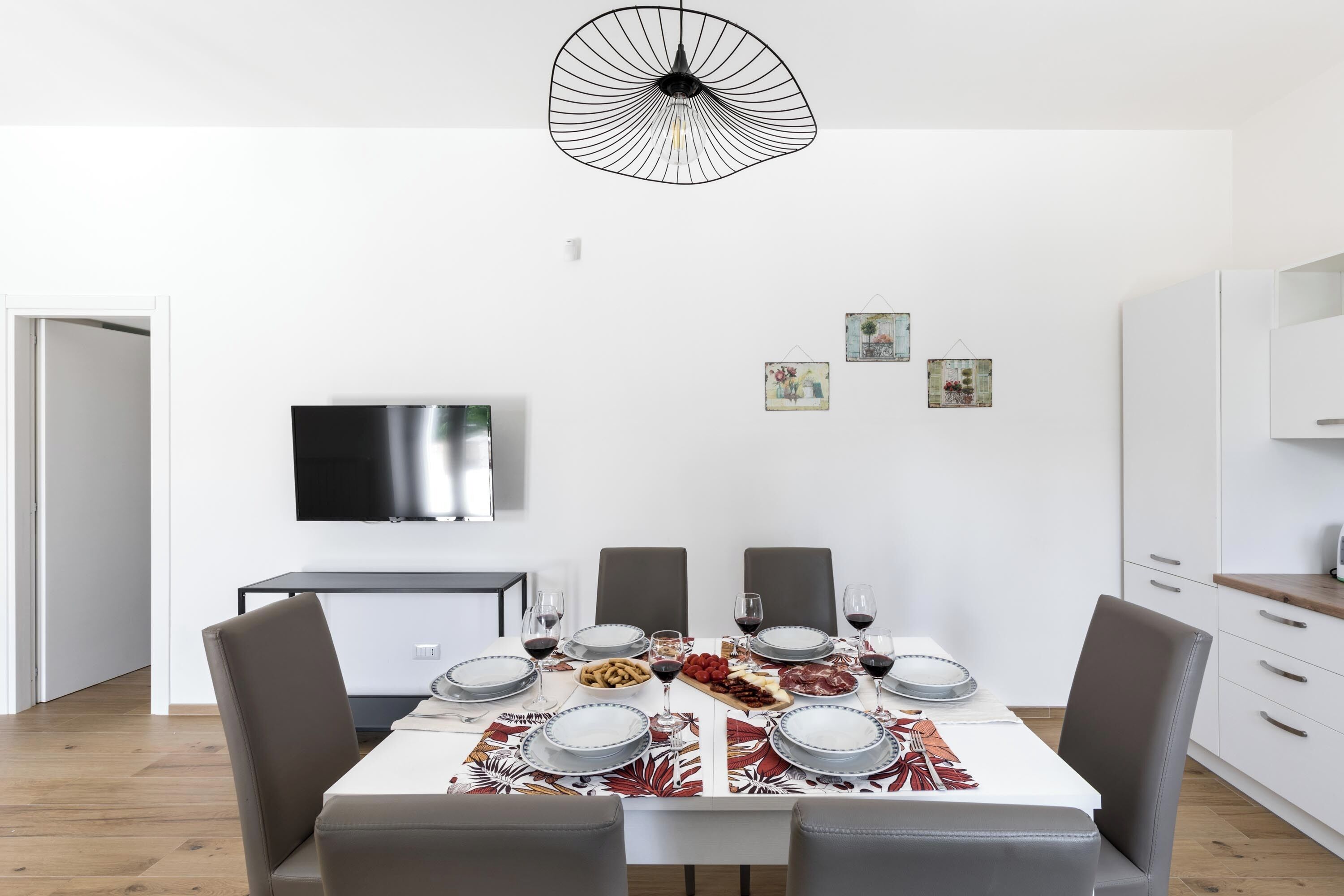 Een moderne eetruimte met een witte tafel gedekt voor vier personen, omringd door grijze stoelen. Daarboven hangt een zwart hanglamp, en aan de muur is een flatscreen televisie bevestigd.