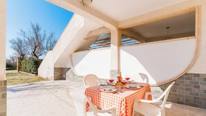 Un espacio de comedor al aire libre cubierto con mantel a cuadros, sillas blancas y vista al cielo y árboles.