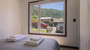 A bedroom with a large window, a bed with two folded towels, and a view of a rural area with cars and buildings. - Tiny House (San Martin de los Andes)