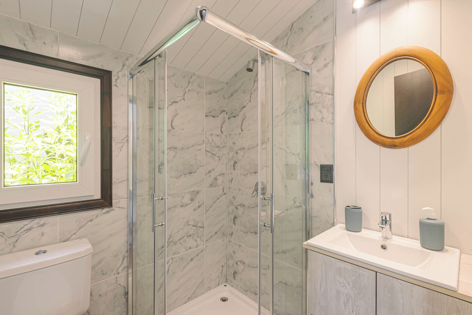 Et badeværelse med en glasafskærmet brusebad, toilet, vask med spejl og et vindue med udsigt til planter.