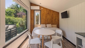 A modern room with a round table and chairs, a bed with wooden paneling, a TV mounted on the wall, and a large window overlooking a garden. - Tiny House (San Martin de los Andes)