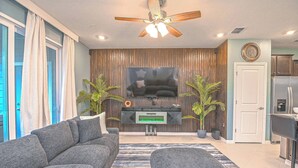 Un salon moderne avec un canapé gris, un pouf gris, un téléviseur à écran plat, un ventilateur de plafond et un coin cuisine équipé d’un réfrigérateur et de placards.