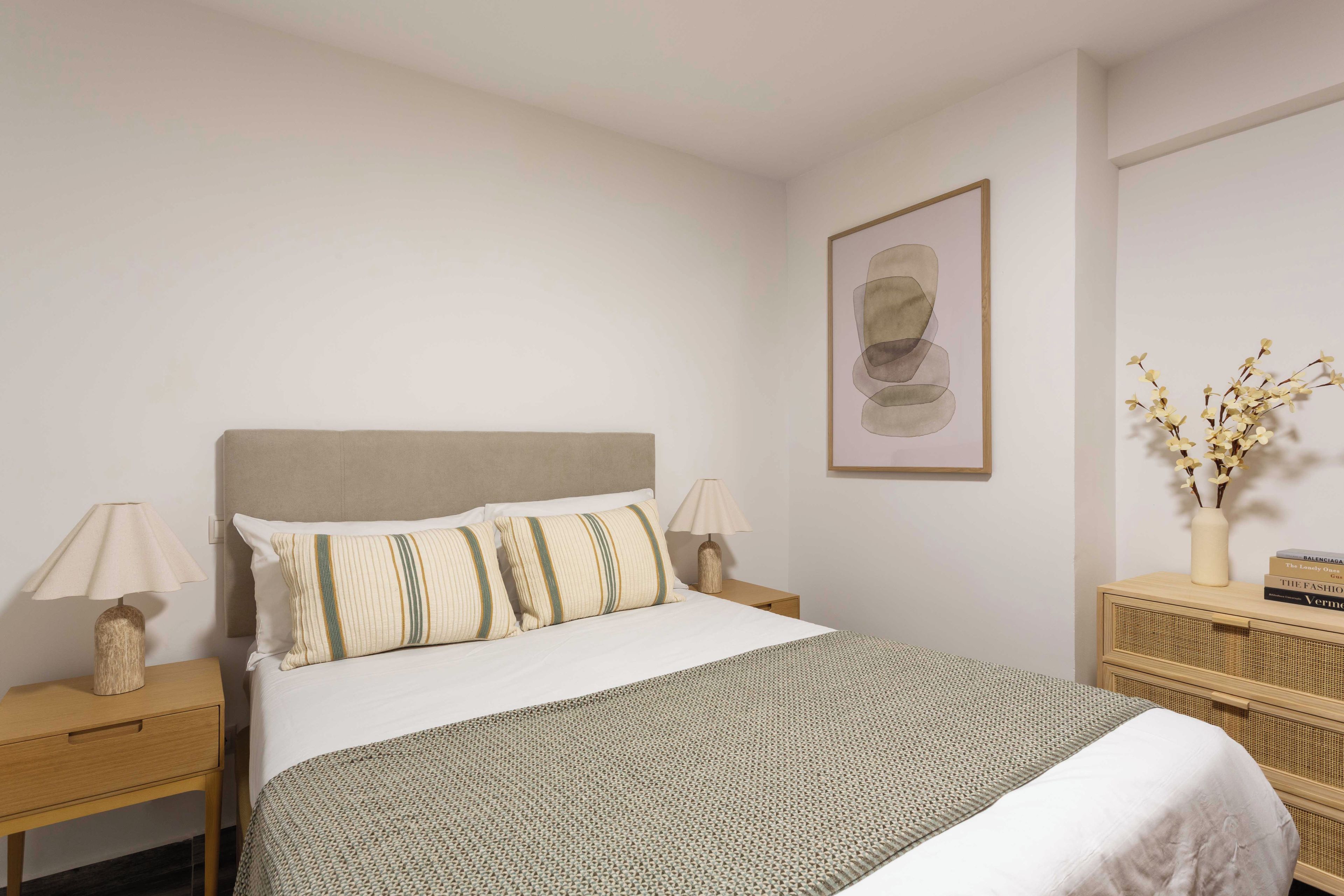 A hotel room with a bed, two bedside tables, a lamp, a framed picture on the wall, and a vase with branches on a side table.