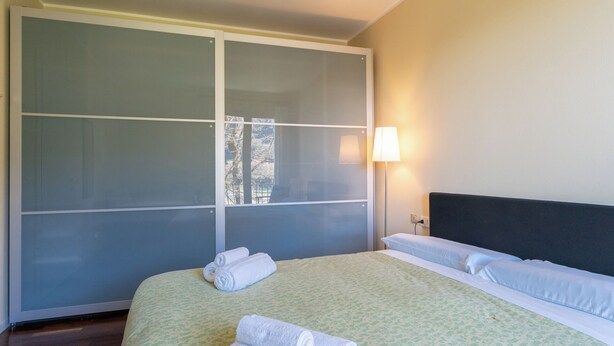 Una habitación de hotel con un amplio ventanal corredero, una cama con sábanas blancas y una lámpara de noche.