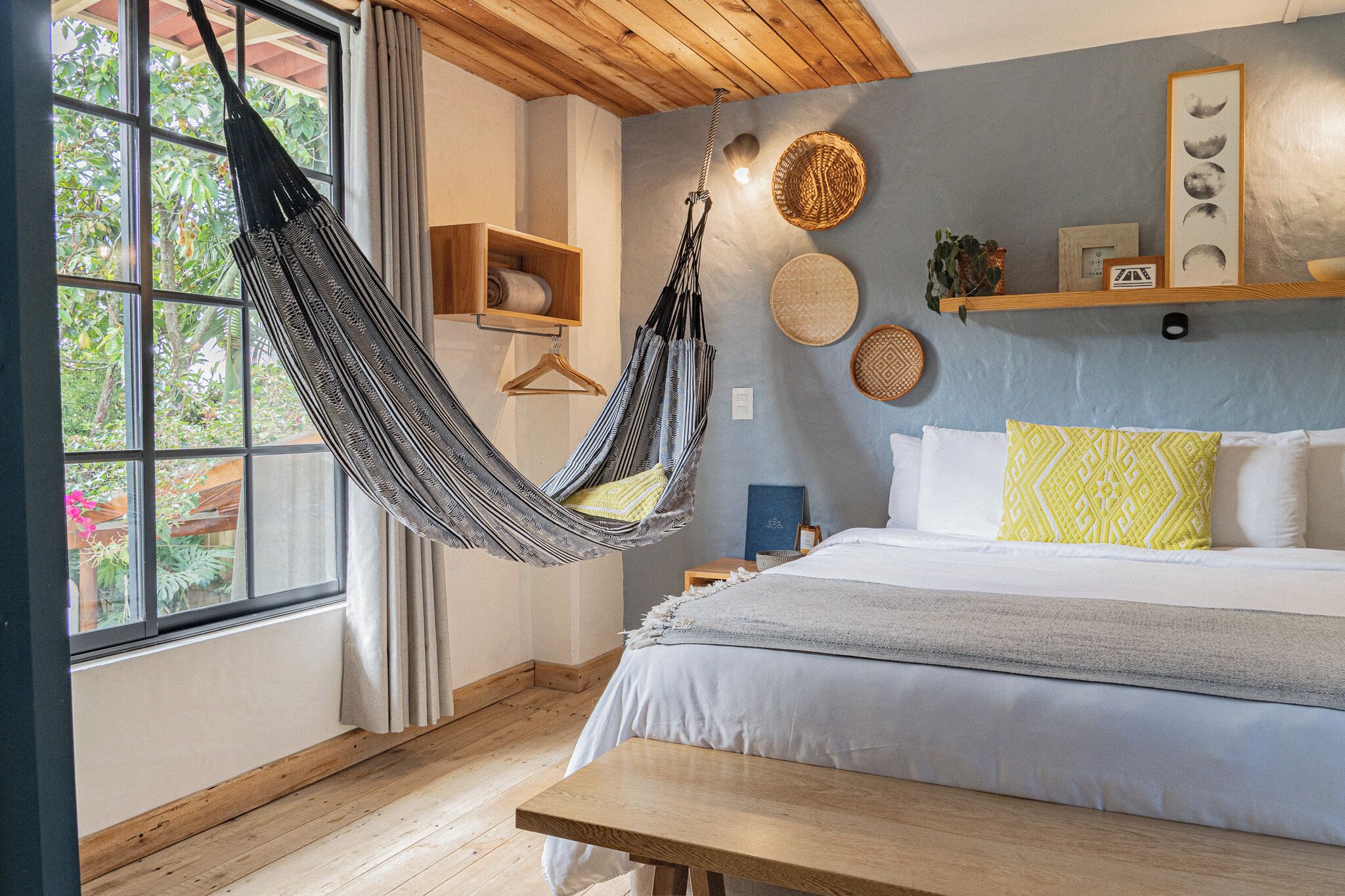 A bedroom with a bed, a hanging hammock, a wooden bench, a window with a view of greenery, and a wall with decorative items.