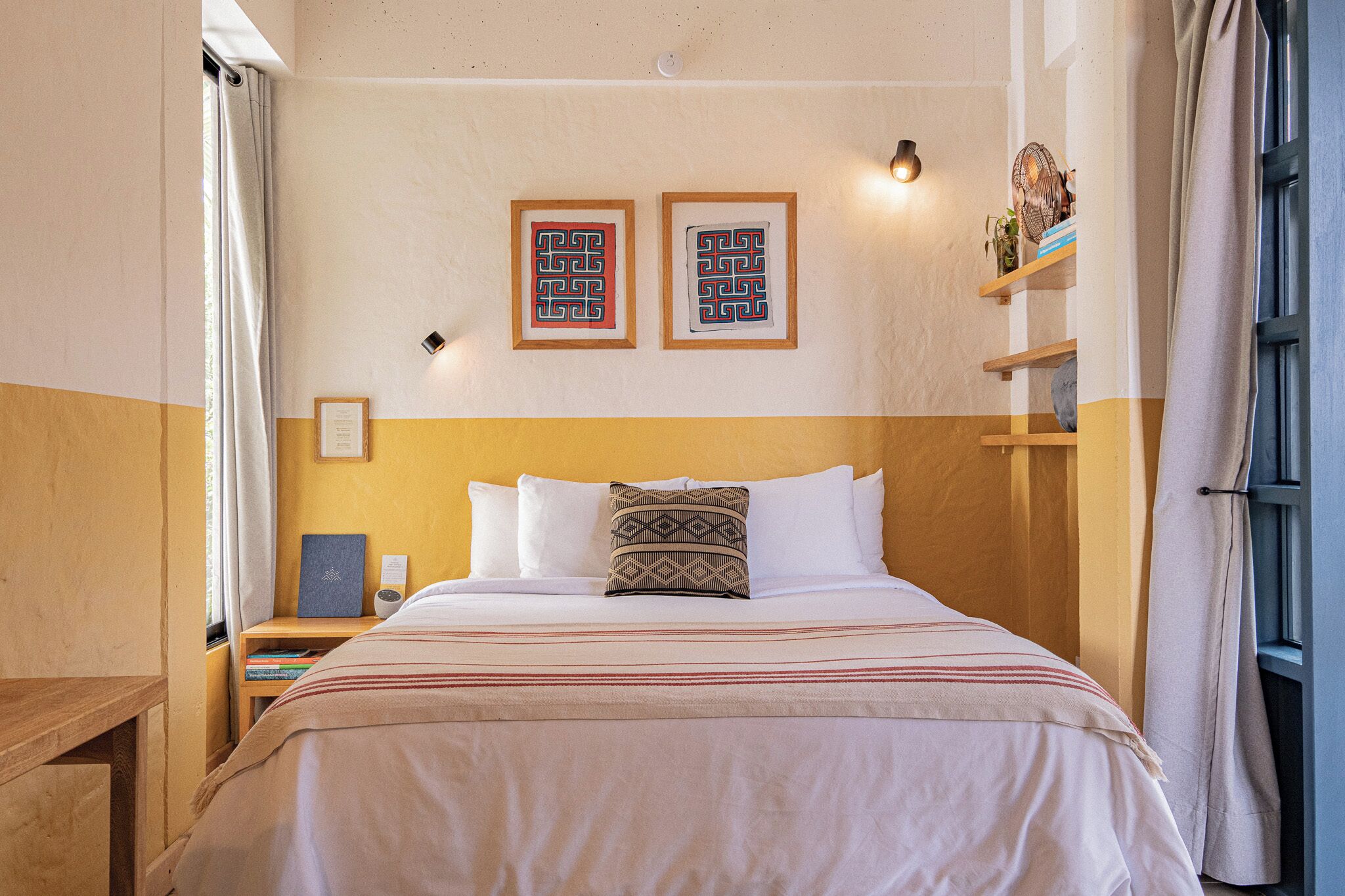 A neatly made bed with a striped blanket, two framed wall art pieces, a bedside table with a lamp, and a shelf with decorative items.