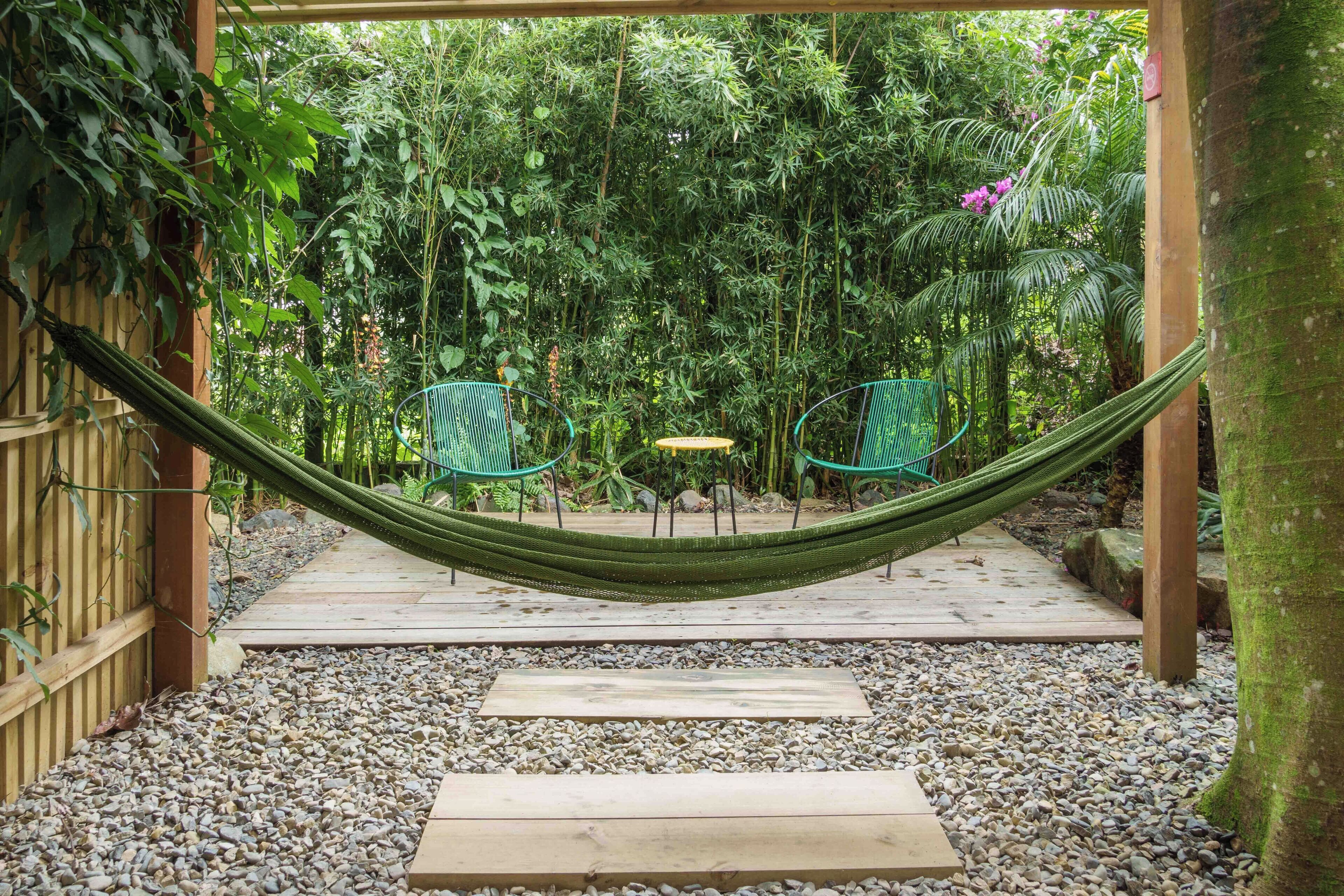 A hammock between two green chairs in a garden with a wooden deck and pebbles.