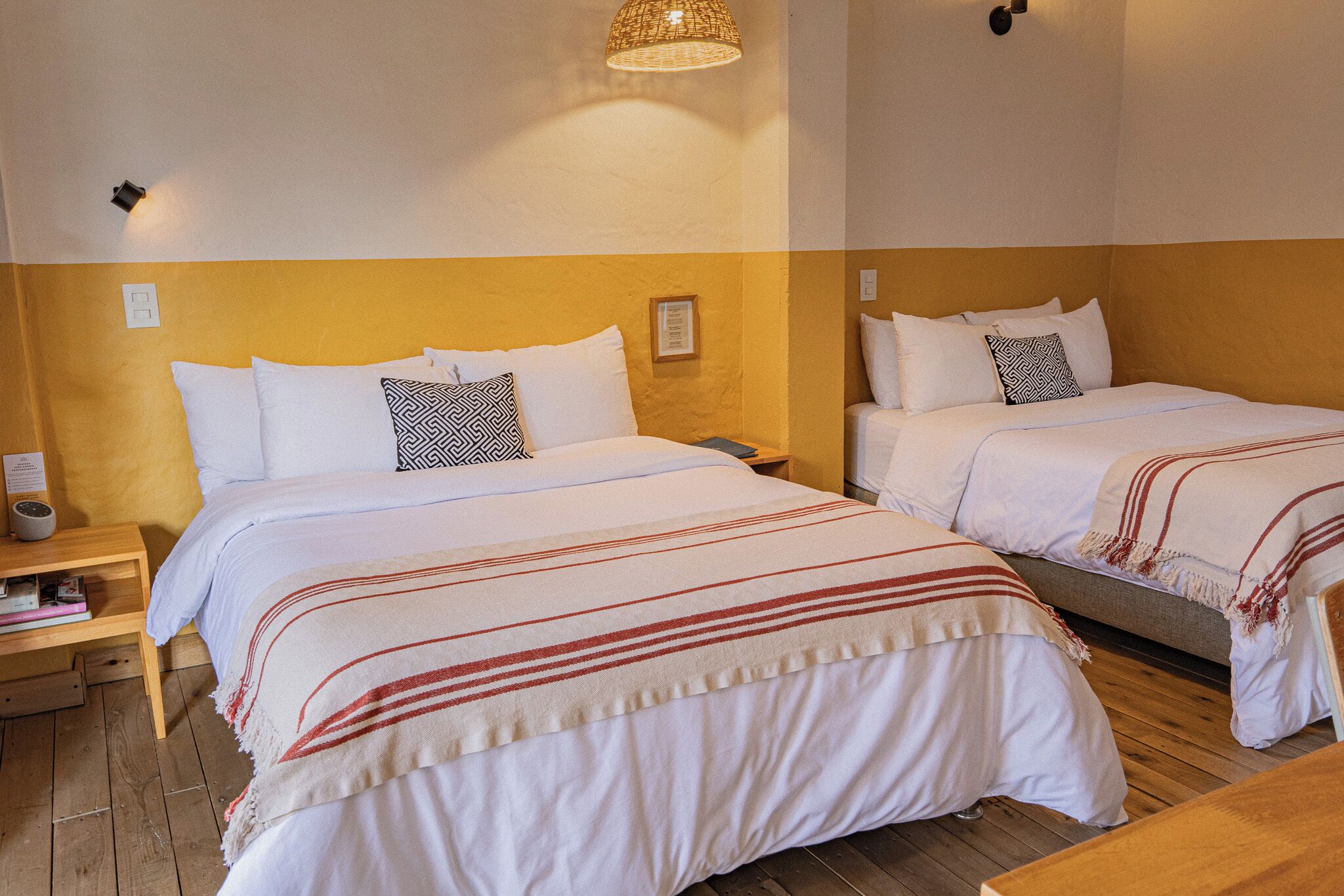 A hotel room with two beds, wooden headboards, a bedside table with a lamp, and a wall-mounted light fixture.