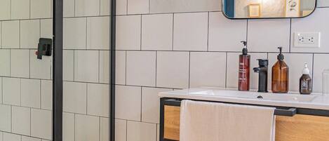 A bathroom with a shower, a sink with toiletries, a mirror, and a towel rack.