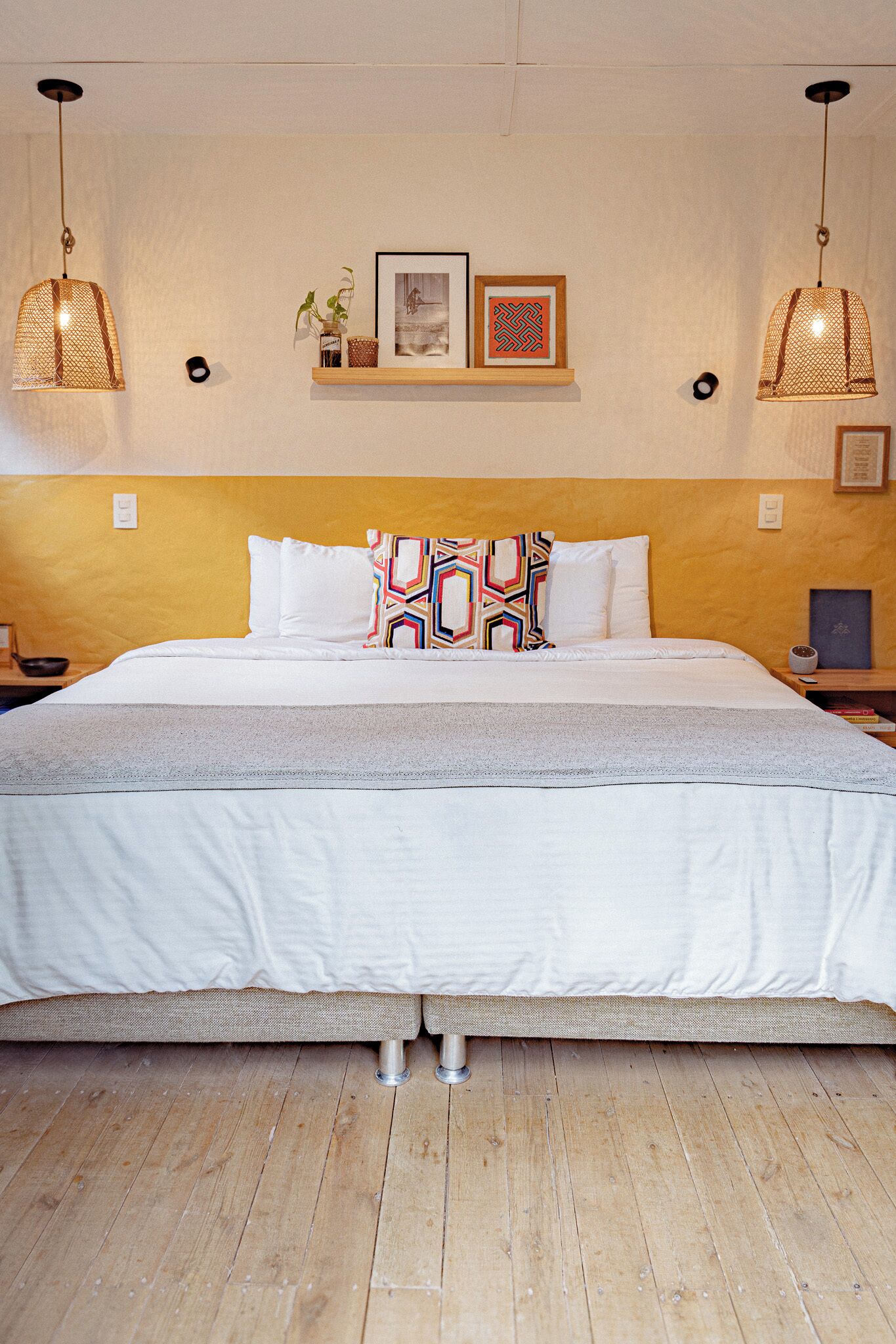 Un dormitorio con una cama, dos mesitas de noche, un estante con fotografías enmarcadas y dos lámparas colgantes.